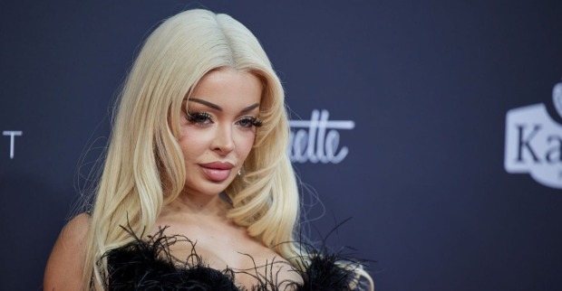 Katja Krasavice mit blondem Haar und tiefem Ausschnitt auf dem roten Teppich, schaut lasziv in die Kamera.