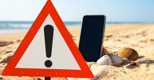 Warnschild und Handy im Sand am Strand: Symbolbild für Kostenfallen durch Handy-Nutzung im Urlaub.
