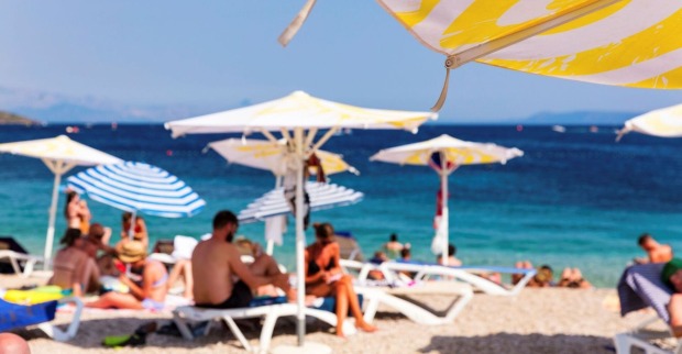 Aufnahme eines mediterranen Strandes mit unscharf zu erkennenden Badegästen, im Vordergrund ein Sonnenschirm