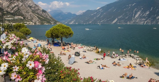 Menschen liegen auf Handtüchern und baden am Strand von Limone sul Garda, mit Blick auf den Gardasee und umliegende Berge, Blumen im Vordergrund