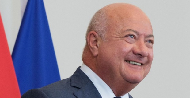 Porträt von Kanzler Christian Stocker, lachend im Anzug, im Hintergrund unscharf rote und blaue Flaggen.