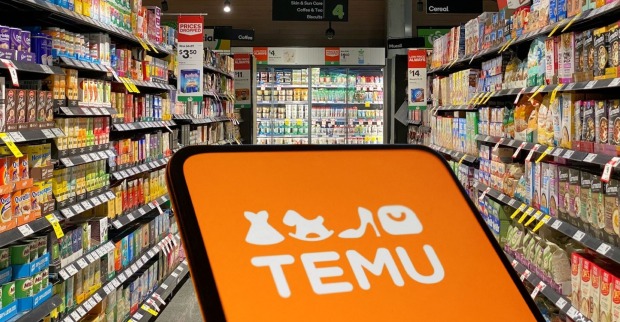 Ein Smartphone mit dem orangefarbenen Temu-Logo im Vordergrund, dahinter ein Supermarktgang mit Regalen voller Lebensmittelpackungen und Kühlregalen.