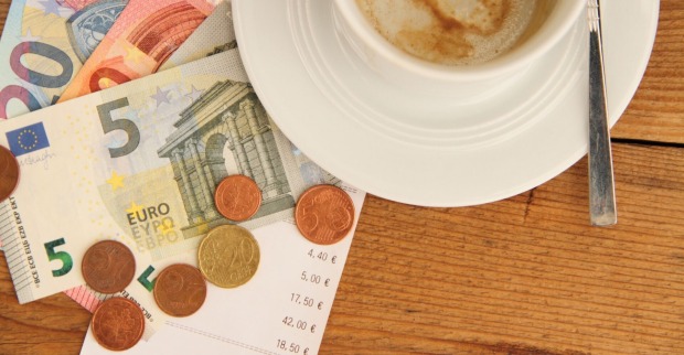 Mehrere Euro-Scheine und Münzen liegen auf einer Restaurantrechnung neben einer weißen Kaffeetasse mit Löffel auf einem Holztisch – Symbolbild für Trinkgeld in der Gastronomie.