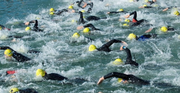Zahlreiche Triathlet:innen mit gelben Badekappen und schwarzen Neoprenanzügen schwimmen eng nebeneinander im offenen Wasser eines Sees. Die Szene zeigt starke Wasserbewegung und Wettkampf-Stimmung.