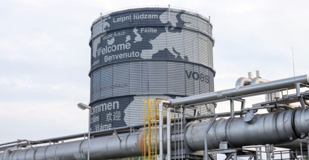Großer, runder Gasspeicher bei der voestalpine in Linz mit internationalen Willkommensgrüßen, umgeben von industriellen Rohrleitungen.