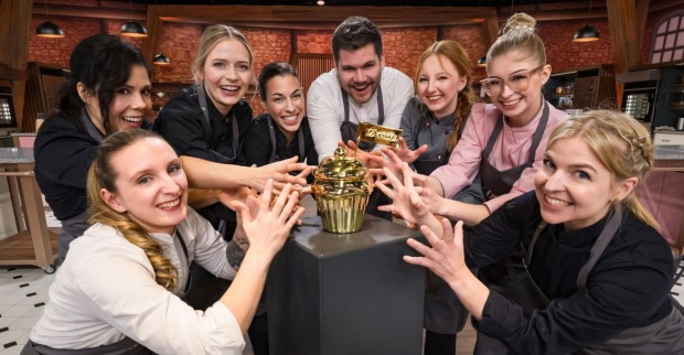 Acht Kandidat:innen von „Das große Backen – Die Profis“ 2025 greifen lachend nach dem Goldenen Cupcake auf einem Podest in der Mitte des Studios.