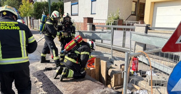 Mehrere Feuerwehrleute in Schutzkleidung und mit Atemschutz arbeiten an einer abgesicherten Baugrube auf einer Straße in Mödling nach einem Gasaustritt.