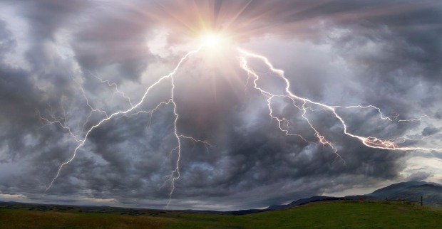 Mehrere Blitze zucken aus einer dunklen Gewitterwolke über eine grüne, hügelige Landschaft. Im Hintergrund sind Berge und ein dramatischer Lichtdurchbruch der Sonne sichtbar.