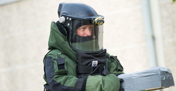 Sprengstoffexperte in schwerer Schutzausrüstung mit Helm und Visier hält ein gesichertes, mit Klebeband umwickeltes Objekt in den Händen.
