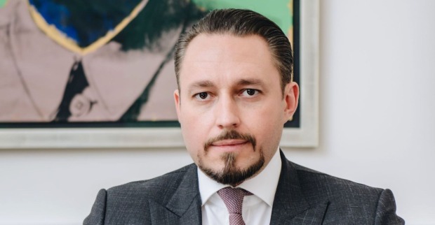 Ein Mann in Anzug und Krawatte blickt direkt in die Kamera. Er hat streng zurückgekämmtes Haar, einen gepflegten Bart und ernsten Gesichtsausdruck. Im Hintergrund ist ein modernes Gemälde zu sehen.