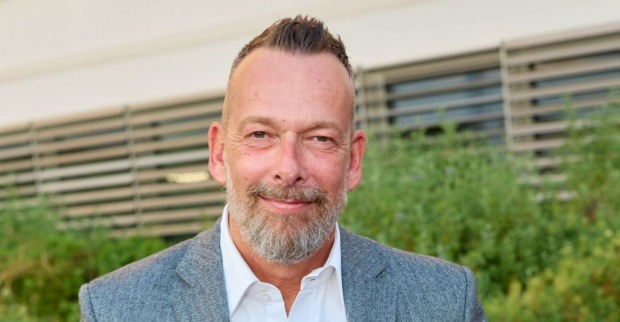 Porträt von Robert Kratky, der im grauen Sakko und weißem Hemd vor einem modernen Gebäude mit Lamellenfassade steht und in die Kamera lächelt.