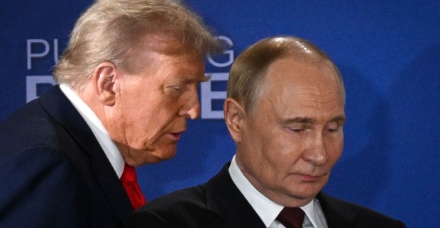 Donald Trump spricht dicht neben Wladimir Putin, der mit ernster Miene nach unten blickt. Beide tragen Anzug, im Hintergrund eine blaue Wand mit dem Schriftzug "Pursuing Peace".