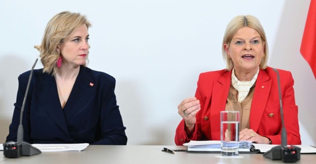 Beate Meinl-Reisinger und Klaudia Tanner sitzen nebeneinander bei einer Pressekonferenz, sprechen vor Mikrofonen, im Hintergrund eine weiße Wand und rot-weiße Fahne.