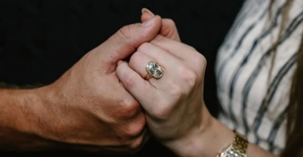 Nahaufnahme zweier sich haltender Hände, im Fokus ein großer rechteckiger Diamantring mit Gelbgoldfassung am Finger von Taylor Swift, die ein gestreiftes Kleid trägt.