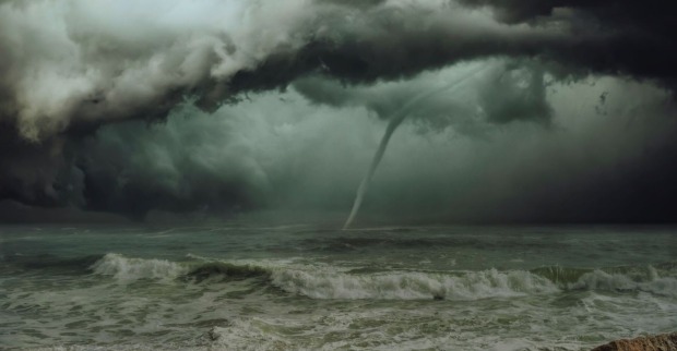 Dunkle Gewitterwolken über dem Meer mit einem gut sichtbaren Tornado, der sich aus der Wolkendecke bis zur Wasseroberfläche erstreckt. Im Vordergrund tobt die Brandung.