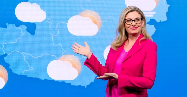 Christa Kummer in pinkem Blazer vor einer Wetterkarte mit Wolken-Symbolen – sie zeigt mit der Hand auf die Grafik und lächelt in die Kamera.
