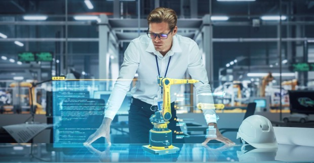 Ein Ingenieur in einem modernen Industriegebäude steht über einem Tisch und betrachtet ein holografisches 3D-Modell eines gelben Roboterarms mit eingeblendeten Programmiercodes und technischen Daten. Neben ihm liegt ein weißer Schutzhelm auf dem Tisch.