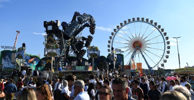 Menschenmenge auf dem Oktoberfest in München, im Hintergrund ein großes Riesenrad und Fahrgeschäfte bei blauem Himmel. Leiche hinter dem Riesenrad auf dem Oktoberfest in München entdeckt – Polizei vermutet medizinischen Notfall.