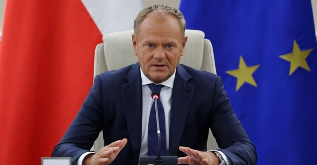 Donald Tusk sitzt in einem dunklen Anzug vor Mikrofonen zwischen einer polnischen und einer EU-Flagge bei einer Krisensitzung.