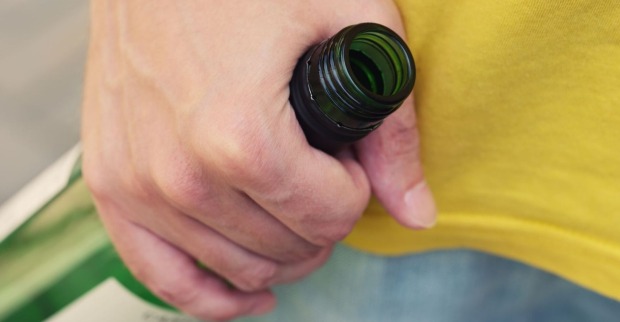 Mann mit gelbem T-Shit hält in aggressiver Grundhaltung Weinflasche aus Grünglas in seiner rechten Hand