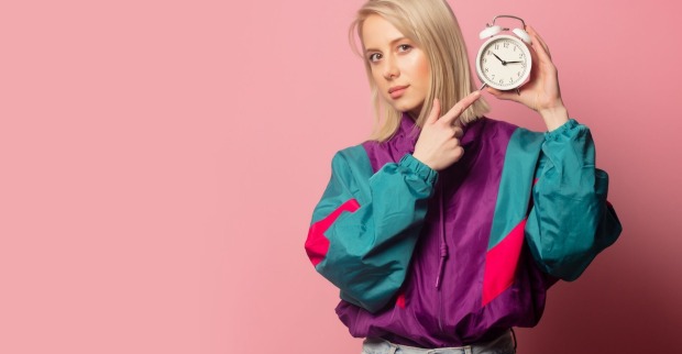 junge blonde Frau in einem bunten Blouson und mit einem klassischen Wecker in der Hand