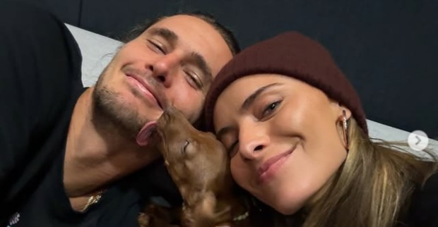 Zverev und Thomalla liegen lächelnd nebeneinander, während ihr kleiner brauner Dackelwelpe ihnen liebevoll über das Gesicht leckt.