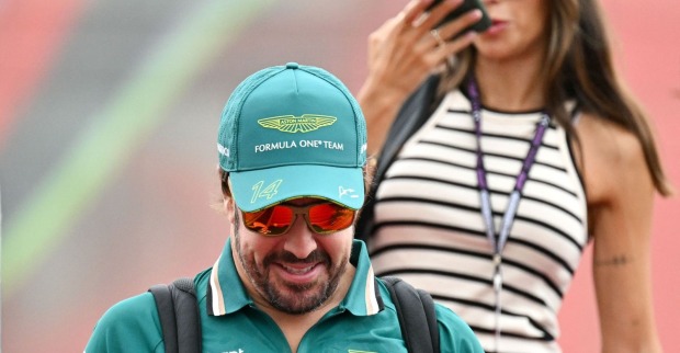 Fernando Alonso in Aston-Martin-Teamkleidung mit Kappe und Sonnenbrille im Paddock, im Hintergrund Melissa Jiménez mit gestreiftem Top und Handy am Ohr. Die beiden erwarten ihr erstes gemeinsames Kind.
