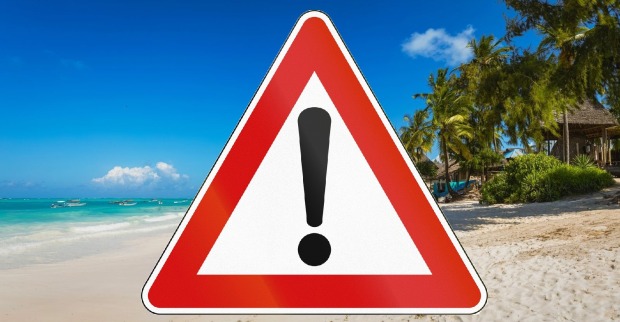 Warnschild mit Ausrufezeichen am tropischen Strand von Tansania als Symbol für aktuelle Reisewarnung.