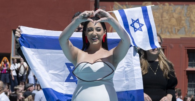 Die israelische ESC-Künstlerin zeigt ein Herz in die Menge, während hinter ihr mehrere Personen israelische Flaggen halten – ein deutliches Zeichen der Unterstützung kurz vor ihrem Auftritt.