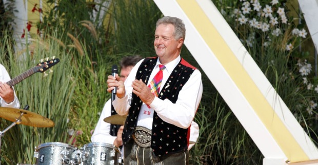 Norbert Rier, Frontsänger der Kastelruther Spatzen, in Tracht hält ein Mikrofon und steht vor einer Band mit Schlagzeug und Gitarre bei einem Auftritt im Freien.