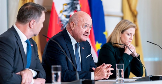 Drei Regierungsmitglieder sitzen bei einer Pressekonferenz vor Mikrofonen und sprechen über die Ergebnisse der Regierungsklausur in Wien. Im Hintergrund sind österreichische und europäische Flaggen zu sehen.