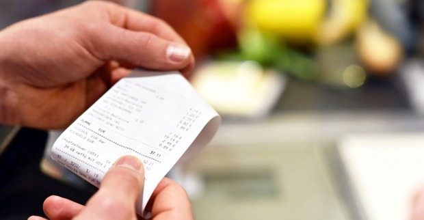 Eine Person hält an der Supermarktkassa einen Kassenbon in der Hand – im Hintergrund sind unscharf Lebensmittel wie Obst und Gemüse zu sehen.