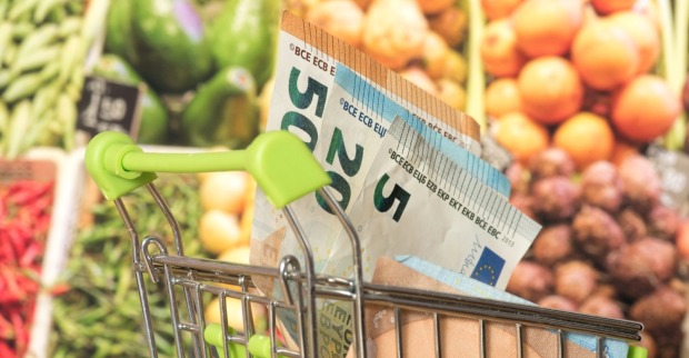 Einkaufswagen mit Euro-Geldscheinen vor einem Gemüseregal im Supermarkt, Symbol für günstigeren Einkauf durch die Mehrwertsteuer-Senkung.