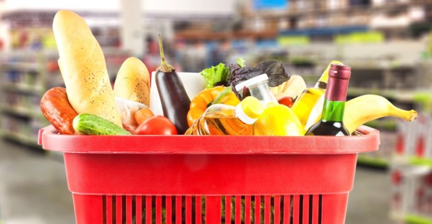 Roter Einkaufskorb mit verschiedenen Lebensmitteln wie Brot, Obst, Gemüse, Öl und Getränken in einem Supermarkt.