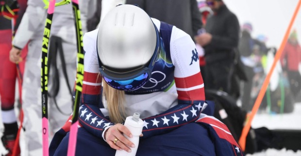 US-Skirennläuferin Lindsey Vonn im Olympia-Rennanzug sitzt erschöpft im Schnee, während das Abfahrtstraining in Cortina wegen Nebel und schwieriger Bedingungen gestoppt ist.