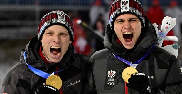 Jan Hörl und Stefan Embacher Teamjacken mit Olympia-Logo halten ihre Goldmedaillen hoch und jubeln nach dem Sieg im Super-Team-Bewerb der Winterspiele 2026.
