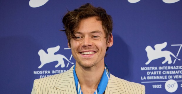 Harry Styles lächelt bei einem öffentlichen Auftritt in hellem Nadelstreifen-Anzug vor blauem Event-Hintergrund.