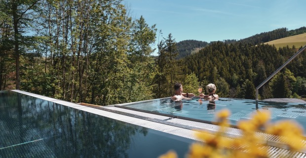 Ein Paar im Outdoor-Pool vor einem Hotel mit Blick auf die Almen | Credit: Hotel Königswieserhof