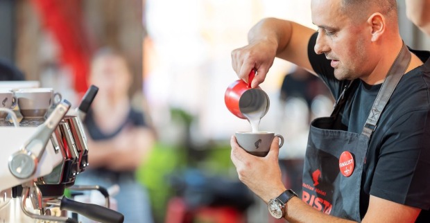 Mann mit Glatze und Schürze füllt vor einer Kaffeemaschine Milchschaum in eine Kaffeetasse