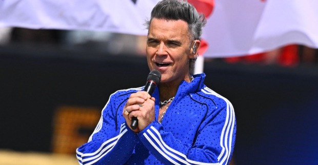 Robbie Williams bei einem Auftritt auf der Bühne im blauen Trainingsanzug mit Mikrofon. Er spricht über seine Autismus-Diagnose