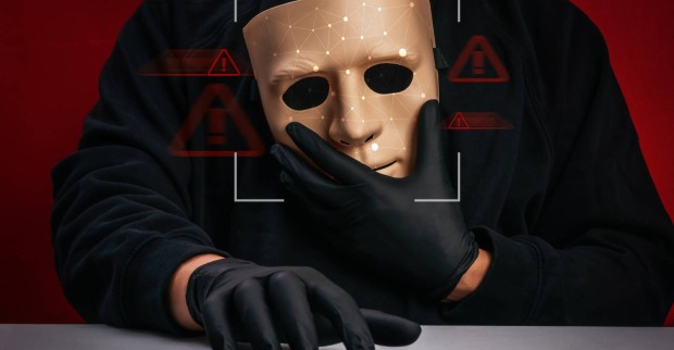 Symbolfoto eines Hackers, der eine Maske in der einen Hand hält und mit der anderen auf einem Keyboard tippt
