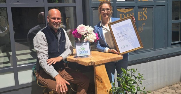 Die Besitzer des Villacher Boutique Hotel Goldenes Lamm, Jakob und Nicole Ottacher, die auf einer Bank vor ihrem Betrieb sitzen und eine Urkunde für die Zertifizierung mit dem EU Ecolabel in die Höhe halten