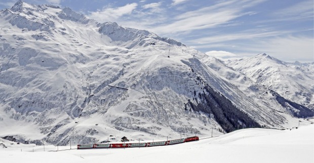 Roter Glacier Express fährt durch eine weitläufige, tief verschneite Berglandschaft in den Schweizer Alpen.
