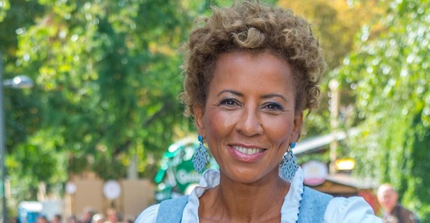 Arabella Kiesbauer lächelt in die Kamera. Die österreichische Moderatorin trägt ein blau-weißes Outfit und auffällige blaue Ohrringe.