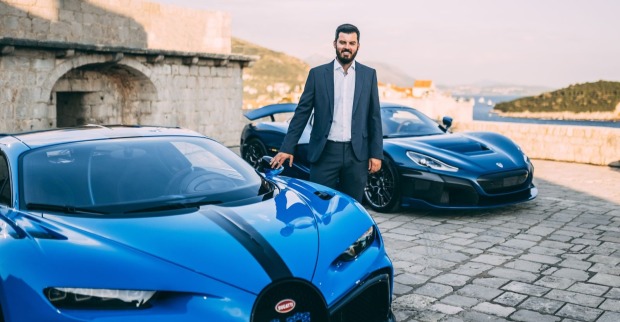 Mate Rimac steht neben einem blauen Bugatti Chiron, im Hintergrund ist ein weiterer blauer Supersportwagen zu sehen.