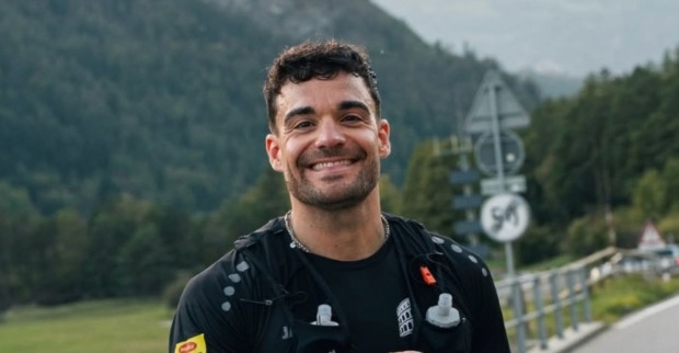 Stefano Zarrella lächelt beim „Run to Rome“ in schwarzer Laufkleidung mit Trinkrucksack auf einer Bergstraße vor bewaldeten Bergen.