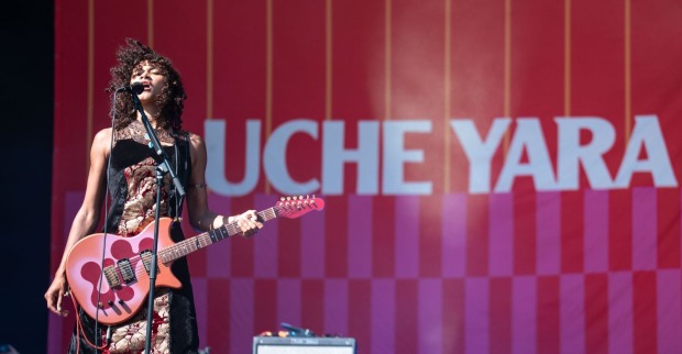 Uche Yara singt bei einem Live-Auftritt in ein Mikrofon und spielt eine rosafarbene E-Gitarre. Im Hintergrund ist ihr Name auf einer rot-pinken Bühnenwand zu sehen.
