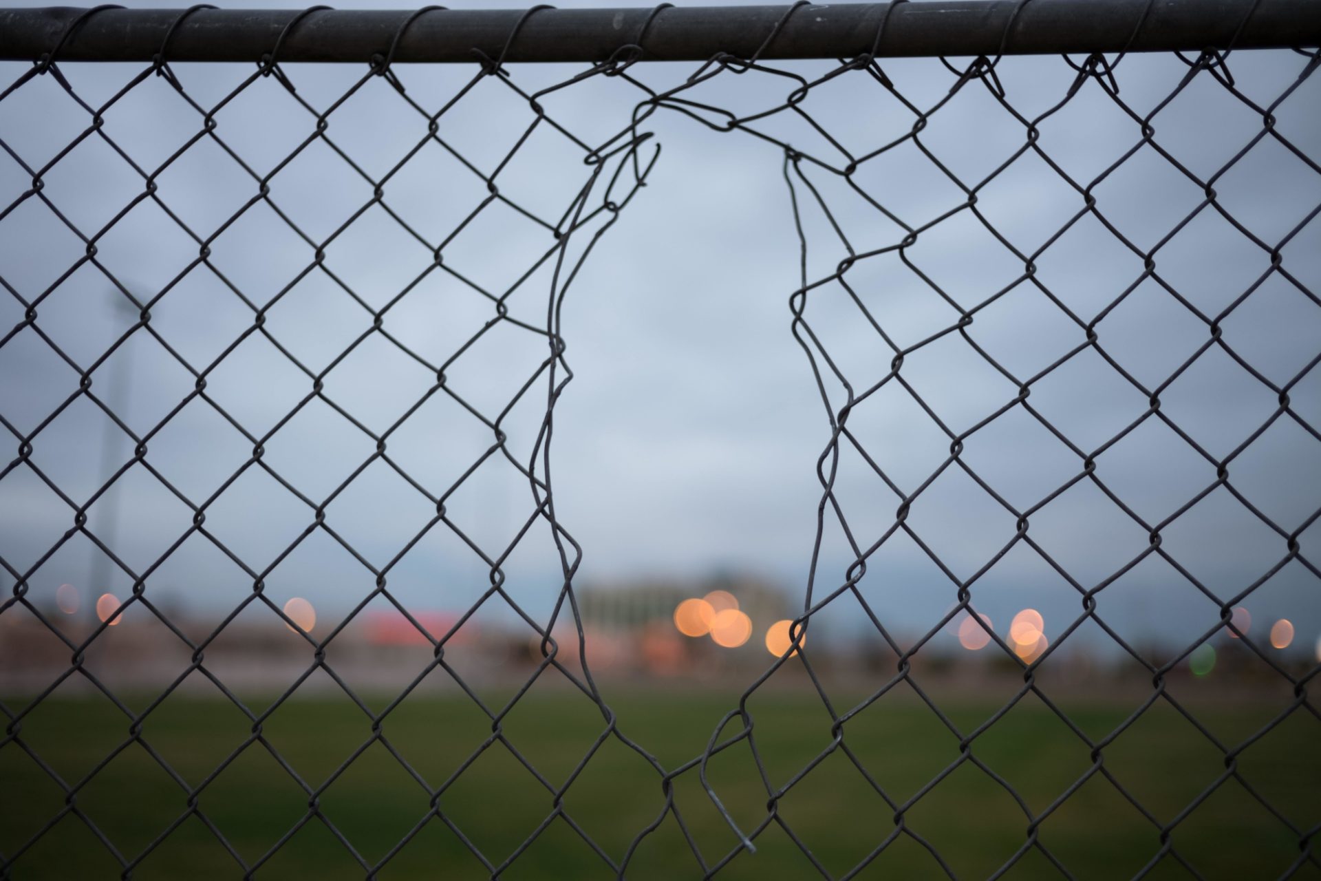 Blick durch das Loch in einem Maschendrahtzaun. In der Ferne sind verschwommen die Lichter von Gebäuden zu sehen.
