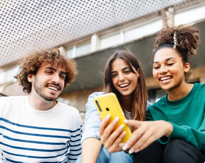 Four international students smiling and sharing a smartphone together outdoors