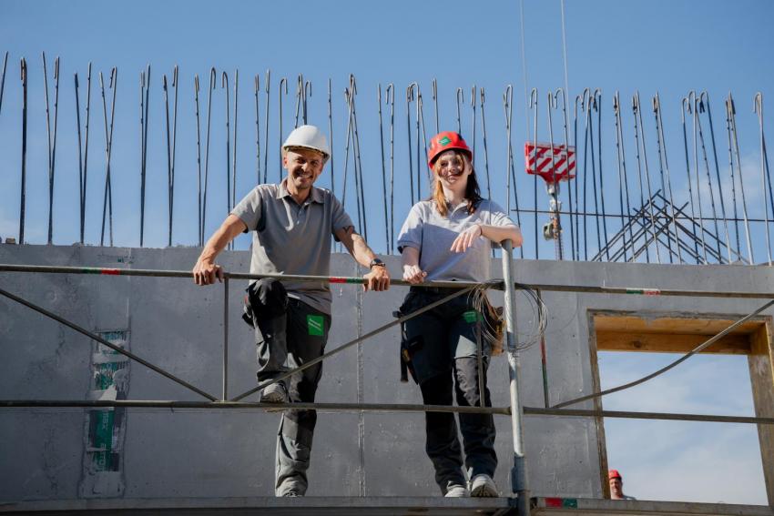 Hochbau-Lehrling Angelina Schadler mit rotem Helm und ihr Ausbildner mit weißem Helm auf einem Baugerüst vor einer Betonwand mit Anschlussbewehrung